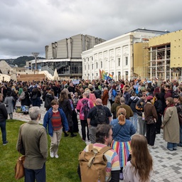 A large crowd of people gathered in a plaza near several buildings.