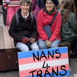 Two women sitting on a park bench with a sign that reads 'NANS 4 TRANS'.