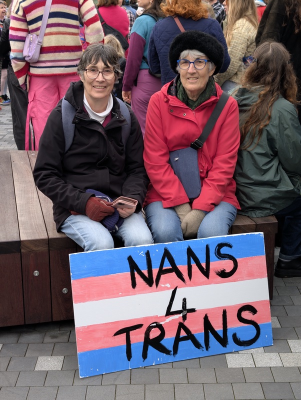Two women sitting on a park bench with a sign that reads 'NANS 4 TRANS'.