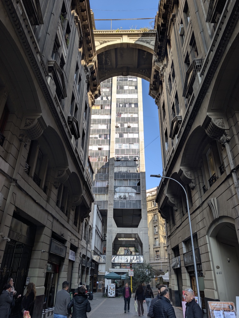 Tall, narrow street flanked by classic architecture and an overhead bridge.